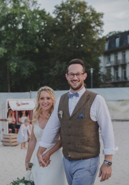 Glückliches Brautpaar schlendert unter freiem Himmel an einem Stadtstrand entlang festgehalten von Hochzeitsfotografin Conny Schöffmann Photography aus Würzburg